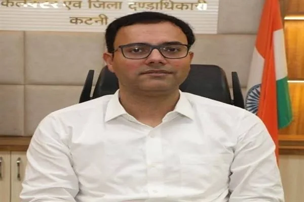 सफेद कमीज और चश्मा पहने व्यक्ति कार्यालय की कुर्सी पर बैठे हैं तथा पीछे भारतीय ध्वज दिखाई दे रहा है